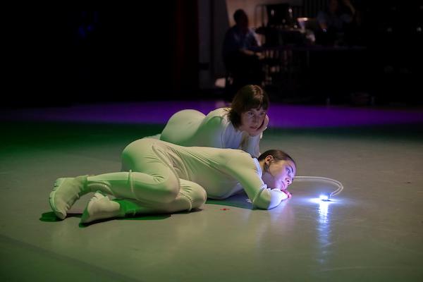 Two dancers in white leotards on the floor interacting with a small moveable robot with a white light on it.