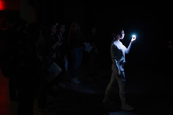 Dancer in white leotard in encompassing black with a small amount of light coming from an interactive robot.